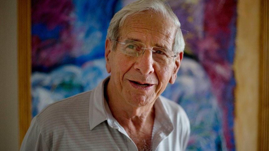 An older man wearing glasses is looking to the left side while smiling. Behind him hangs a colourful painting.