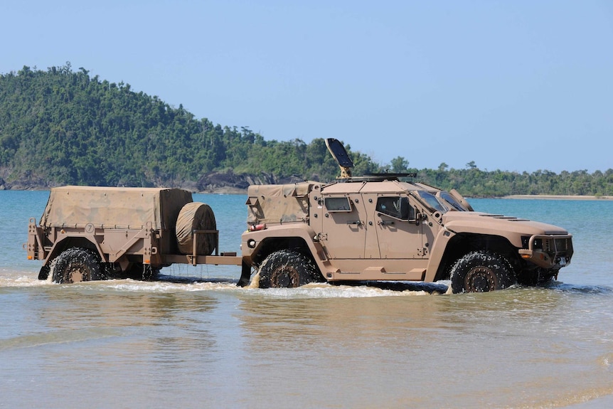 A military four-wheel drive tows a trailer through the shallows at a beach.