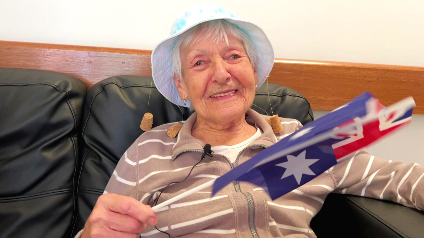 An elderly woman in a hat, smiling at the camera, waving a small Australian flag.