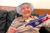 An elderly woman in a hat, smiling at the camera, waving a small Australian flag.