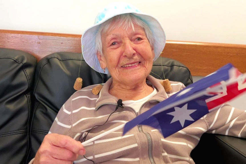 An elderly woman in a hat, smiling at the camera, waving a small Australian flag.