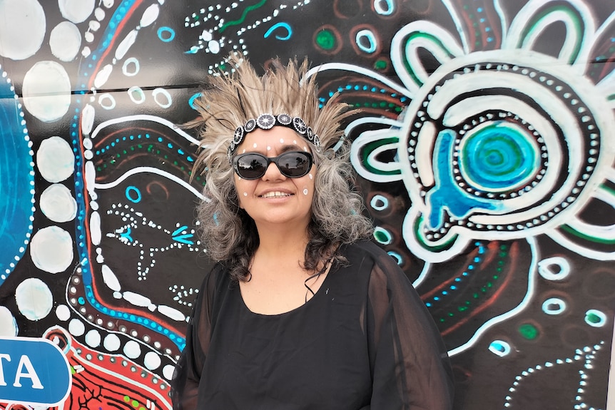 An Aboriginal woman in traditional dress against an indigenous artwork.