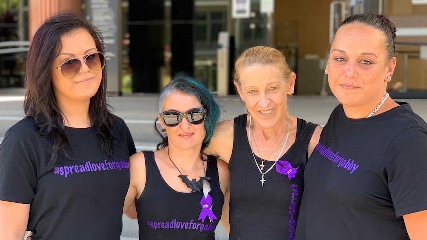 Four women with their arms around each other, all wearing matching blue t-shirts, outside a court house.