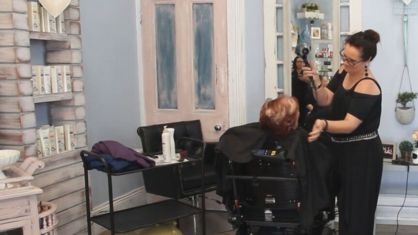 Desiree McDonald stands next to client Betsy White who is in a wheelchair.  Desiree is blow drying Betsy's hair.