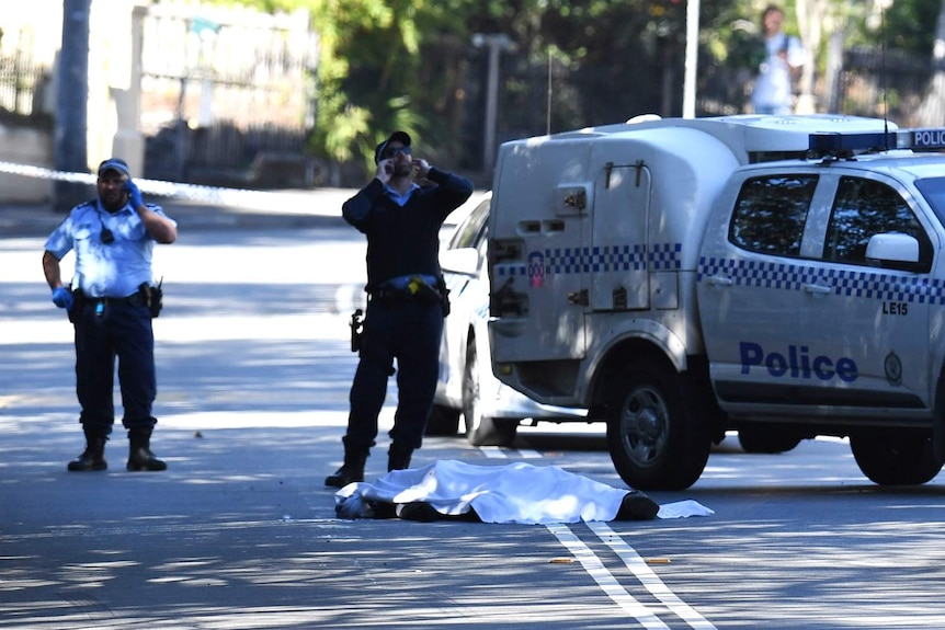 Two men stand near a body, covered in a sheet on the road.