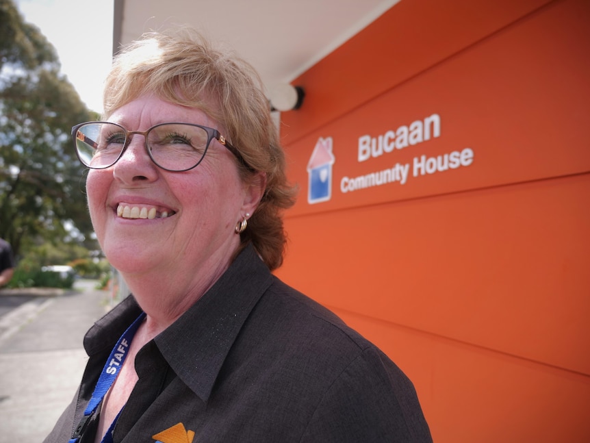 Close shot of a middle aged woman with glasses smiling as she stands in front of a community house.