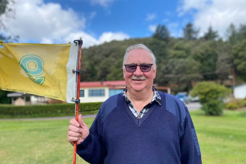 An older man in a blue jumper and checked shirt holds a yellow flag on a golf course.