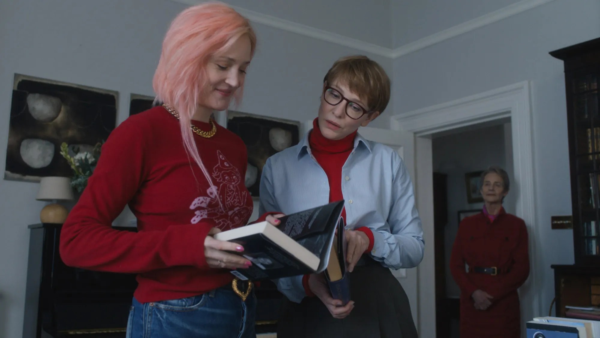 Two women in a stylish room look at a book with their mother in the background  