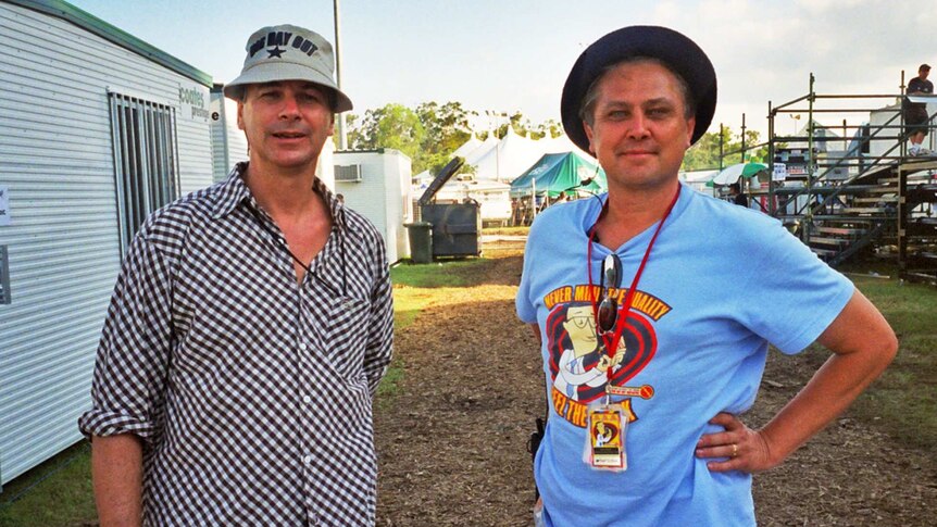 Two men stand between portable buildings at a musical festival.