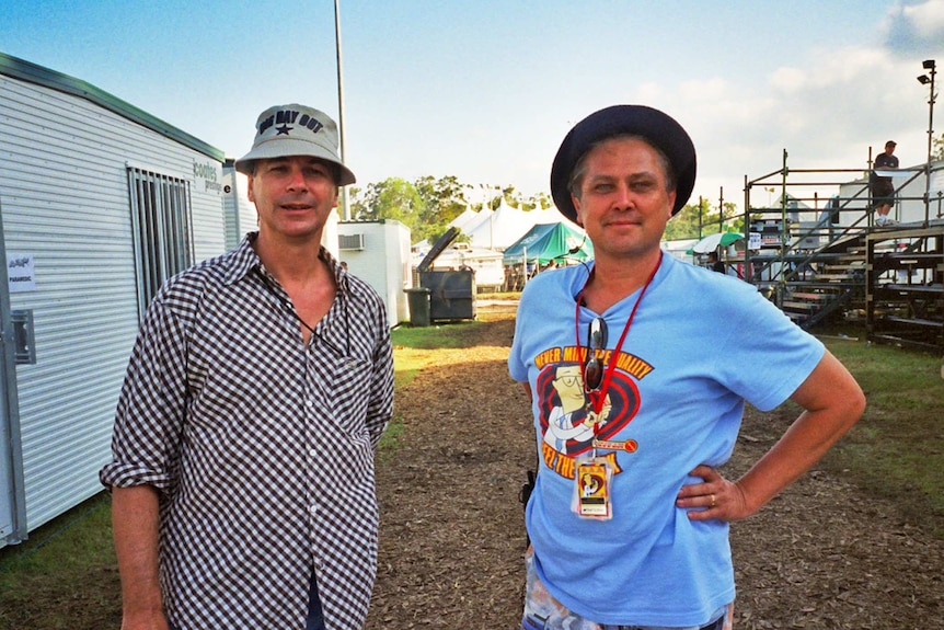 Two men stand between portable buildings at a musical festival.