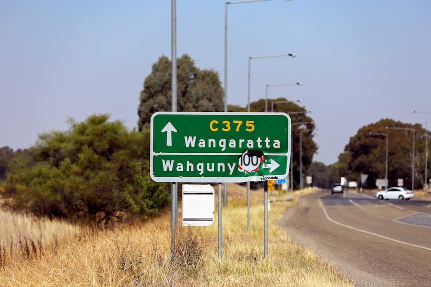 A green road sign next to a country road. There's a large hole in the word Wahgunyah.