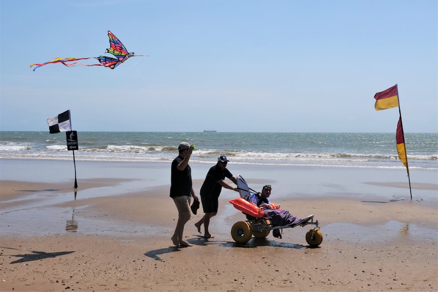 A wide-shot photo of a man being pushed along the beach in a floating wheelchair by a man, while another one flies a kite.