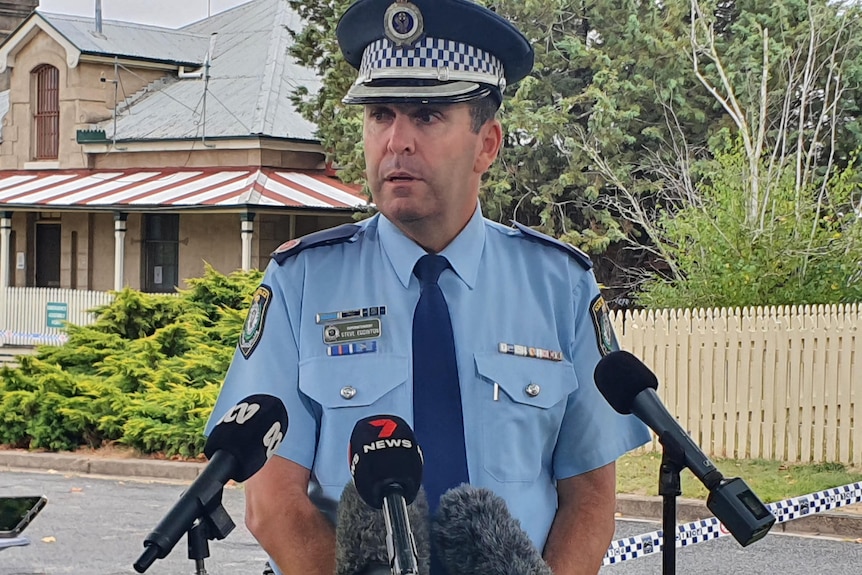A uniformed police officer stands speaking in front of several media outlet microphones, out the front of a police station.
