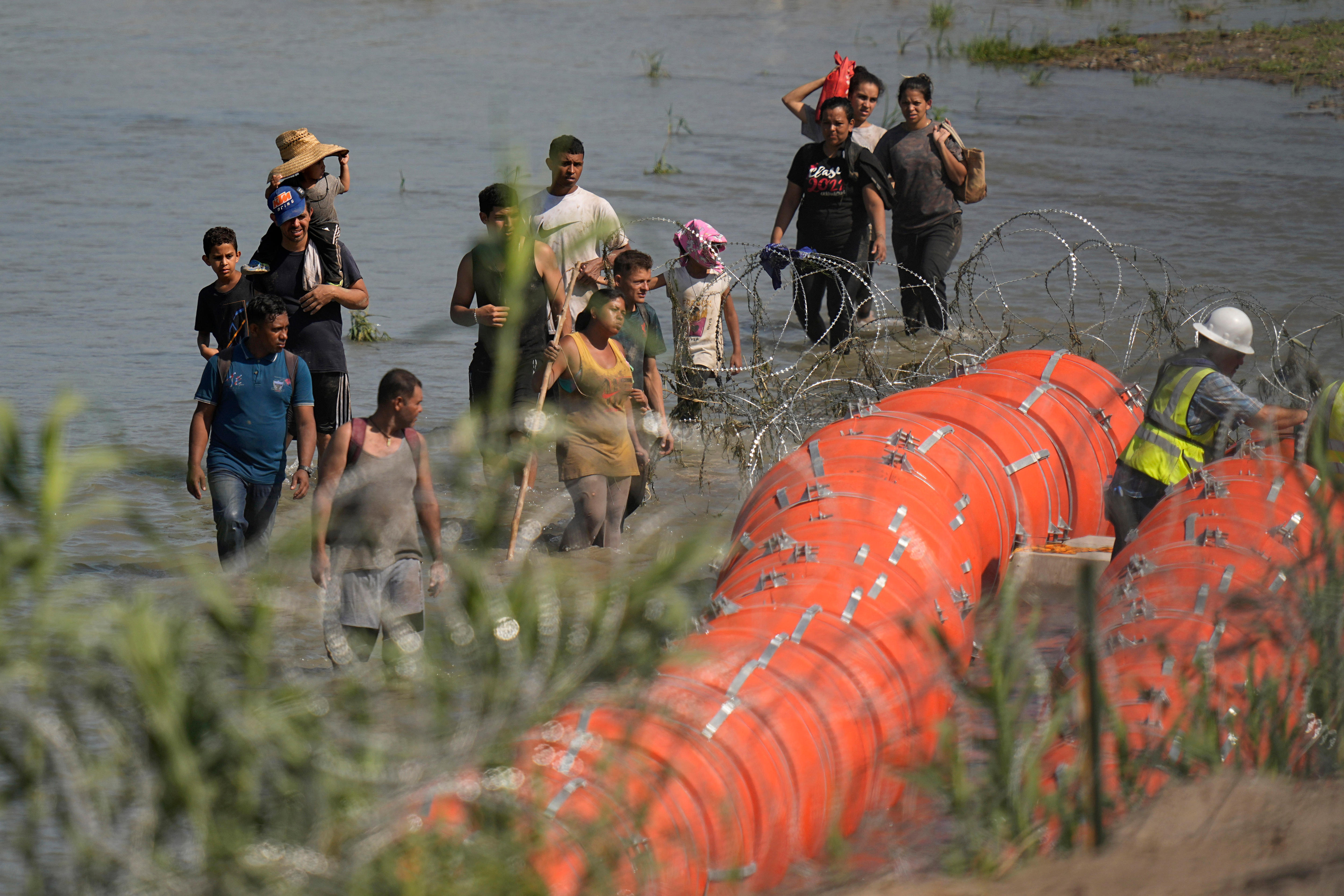 A group of people stand in the water as workers assemble large orange buoys with wire.