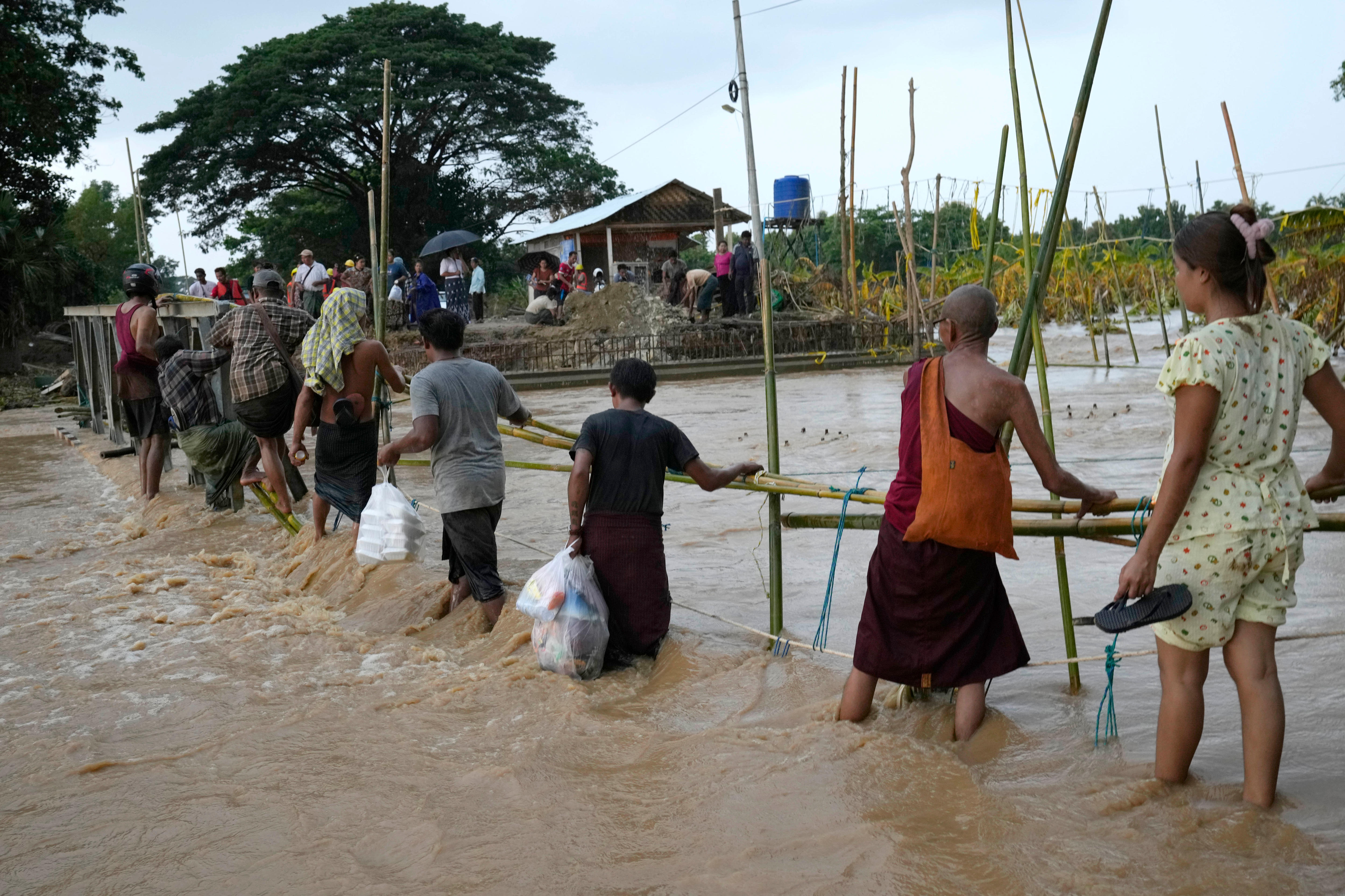 Eight people holding bags and shoes move through floodwaters that are raging across the top of a broken bridge