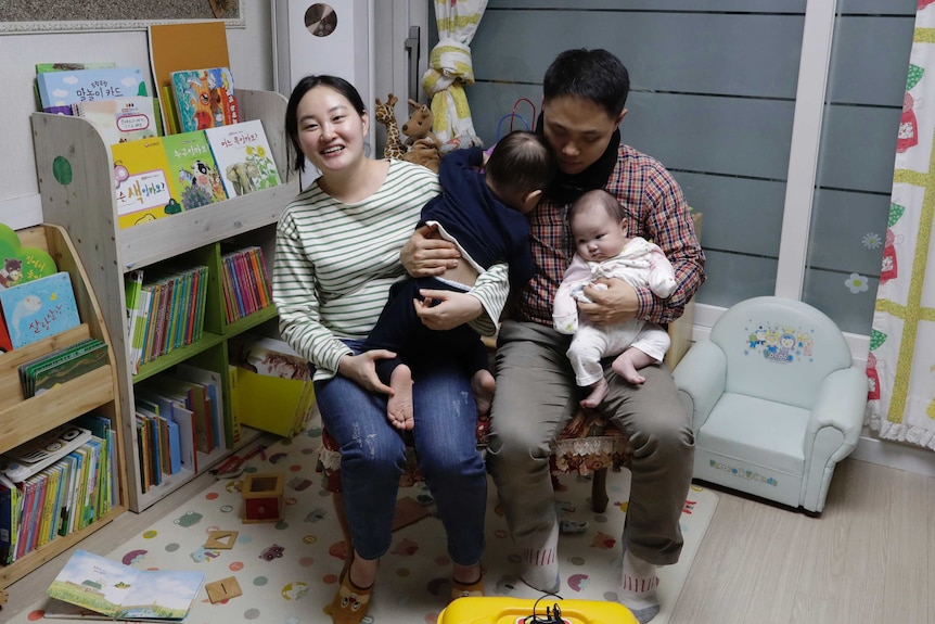 A woman and man hold their two children in a room with books and children's toys.