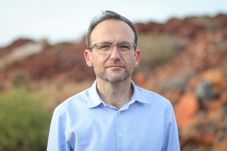 A close-up of a man wearing glasses, standing in front of green and red hills.