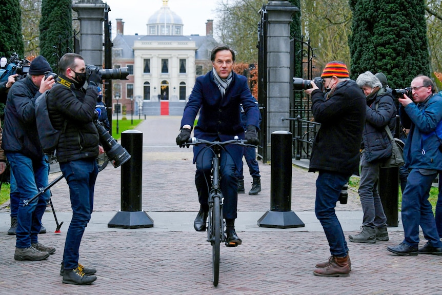 Dutch Prime Minister Mark Rutte riding a bicycle out palace gates surrounded by photographers