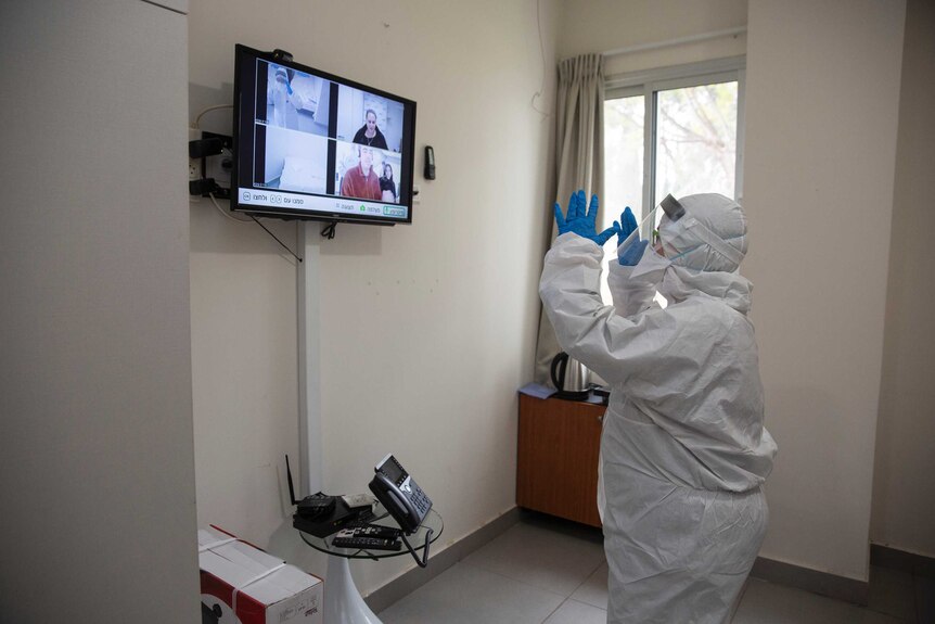 A person dressed in protective clothing, gloves and mask stands in a closed room with a TV screen.