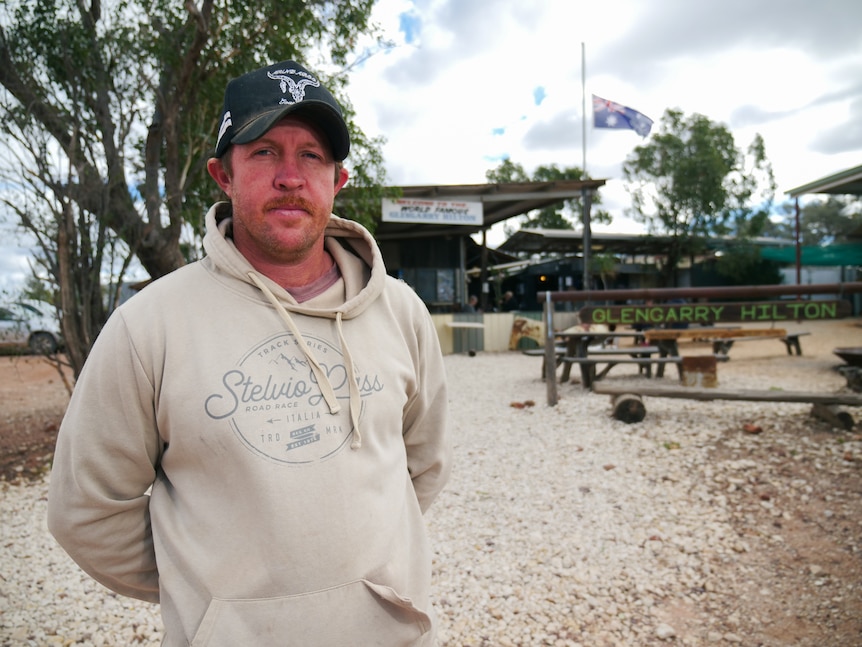 Man stands in grey pullover jumper and cap in front of outback pub.