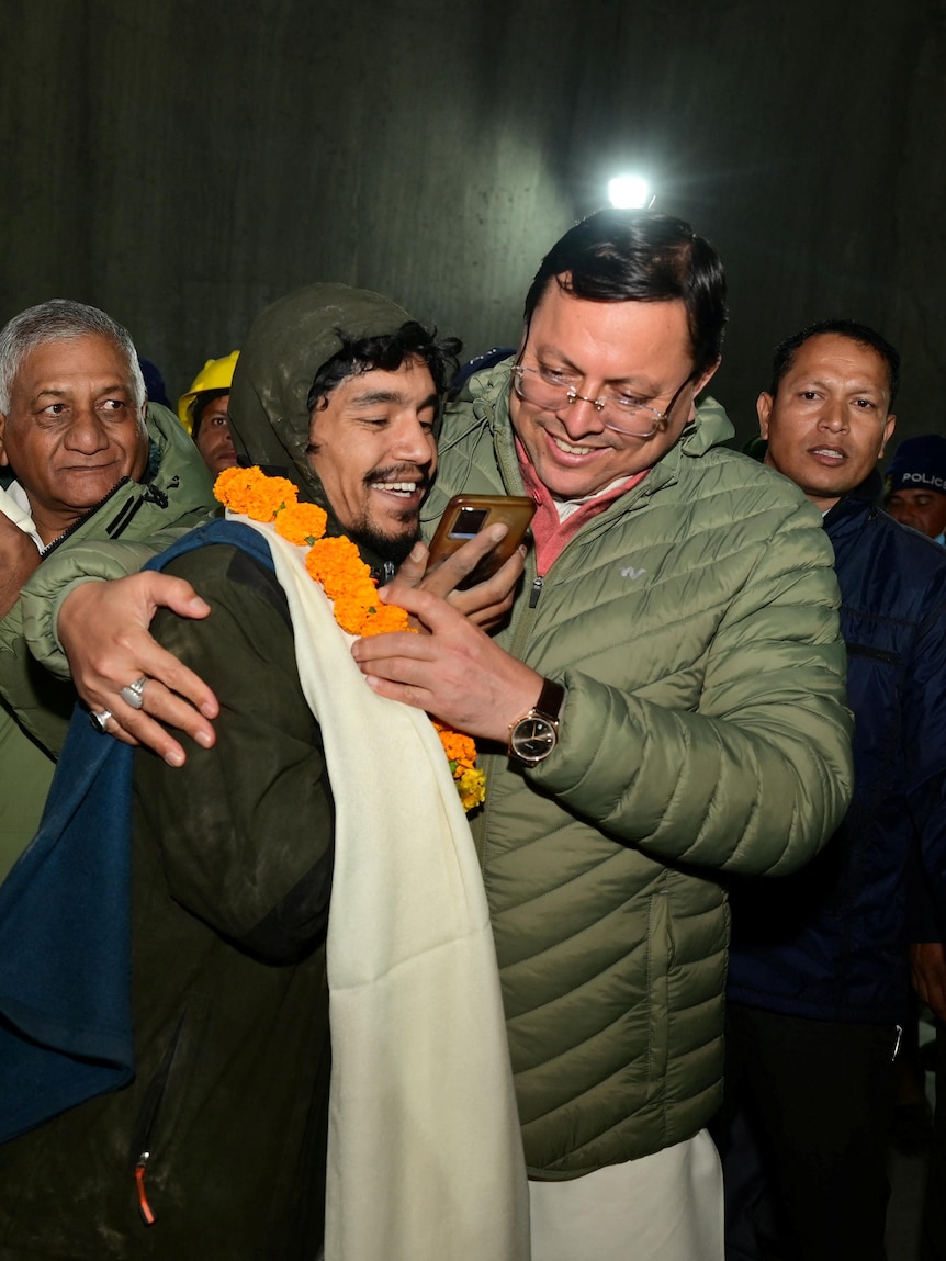 A man wrapped in a jacket and blanket with a flower garland around his neck holding a phone and being hugged by another man