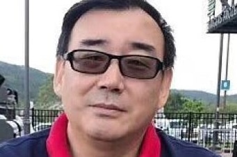 A photo of a Chinese-Australian man Yang Hengjun, sitting on an outside table, wearing a navy polo shirt with a red collar.