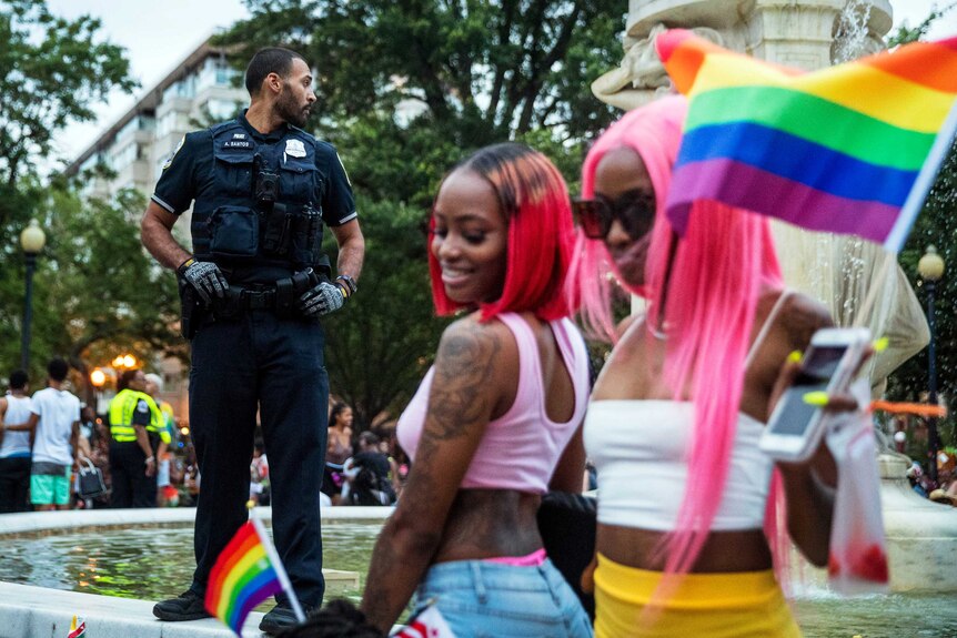 Police stand guard in the background with women wearing pink hair and carrying rainbow flags in the foreground.
