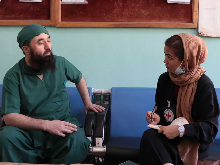 A woman on the right in a head scarf and wearing a red cross pin speaks to a surgeon on the left wearing scrubs