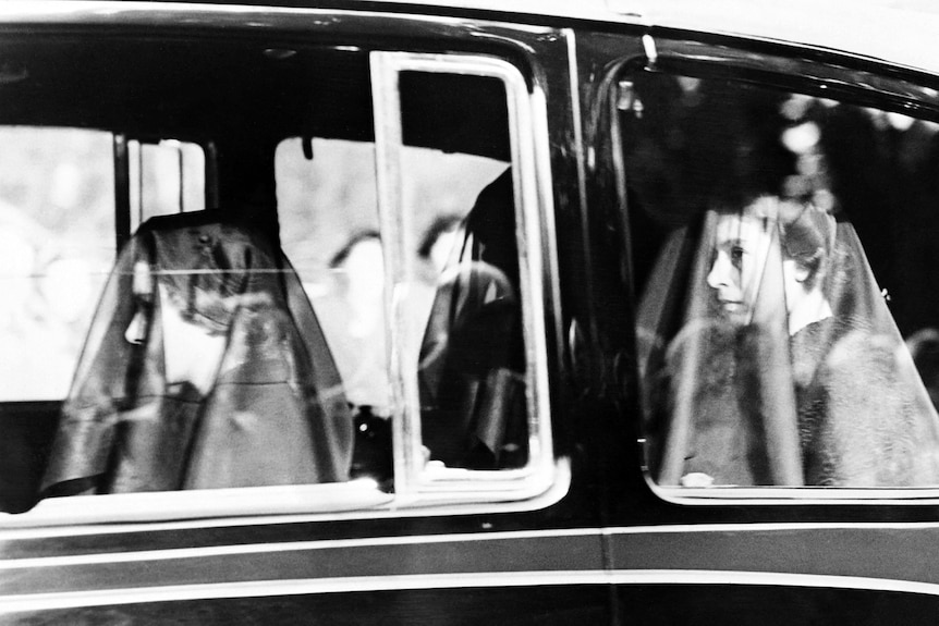A B&W photo of three women in see-through veils travel in a car, looking sombre