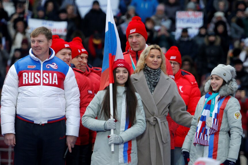 Una atleta rusa sostiene una bandera rusa y sonríe junto a otros atletas en un mitin por Vladimir Putin.