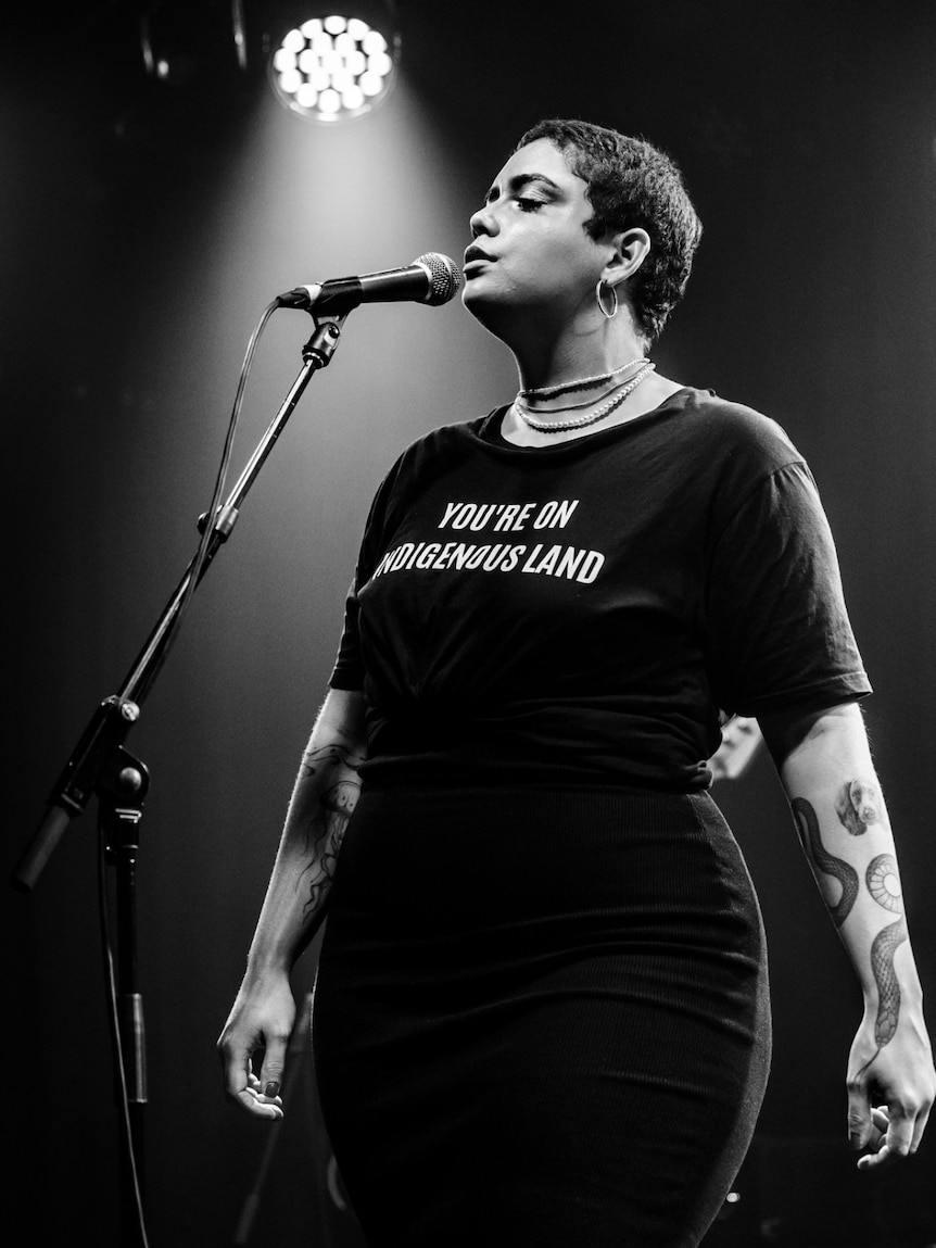 Kee'Ahn singing on a stage, wearing a black t-shirt that reads "You're on Indigenous Land".