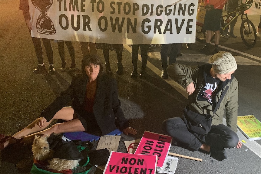 Two people with their hands glued to the road with protesters holding signs behind them.