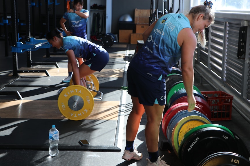 Female rugby players prepare to lift weights in a gym.