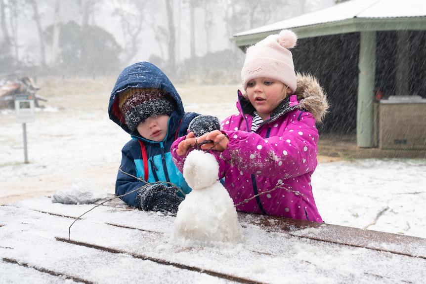 two children in colorful jackets building a snowman on a picnic table