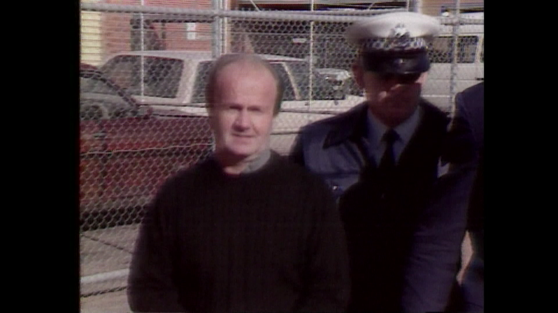 A balding man walks next to a uniformed police officer. 