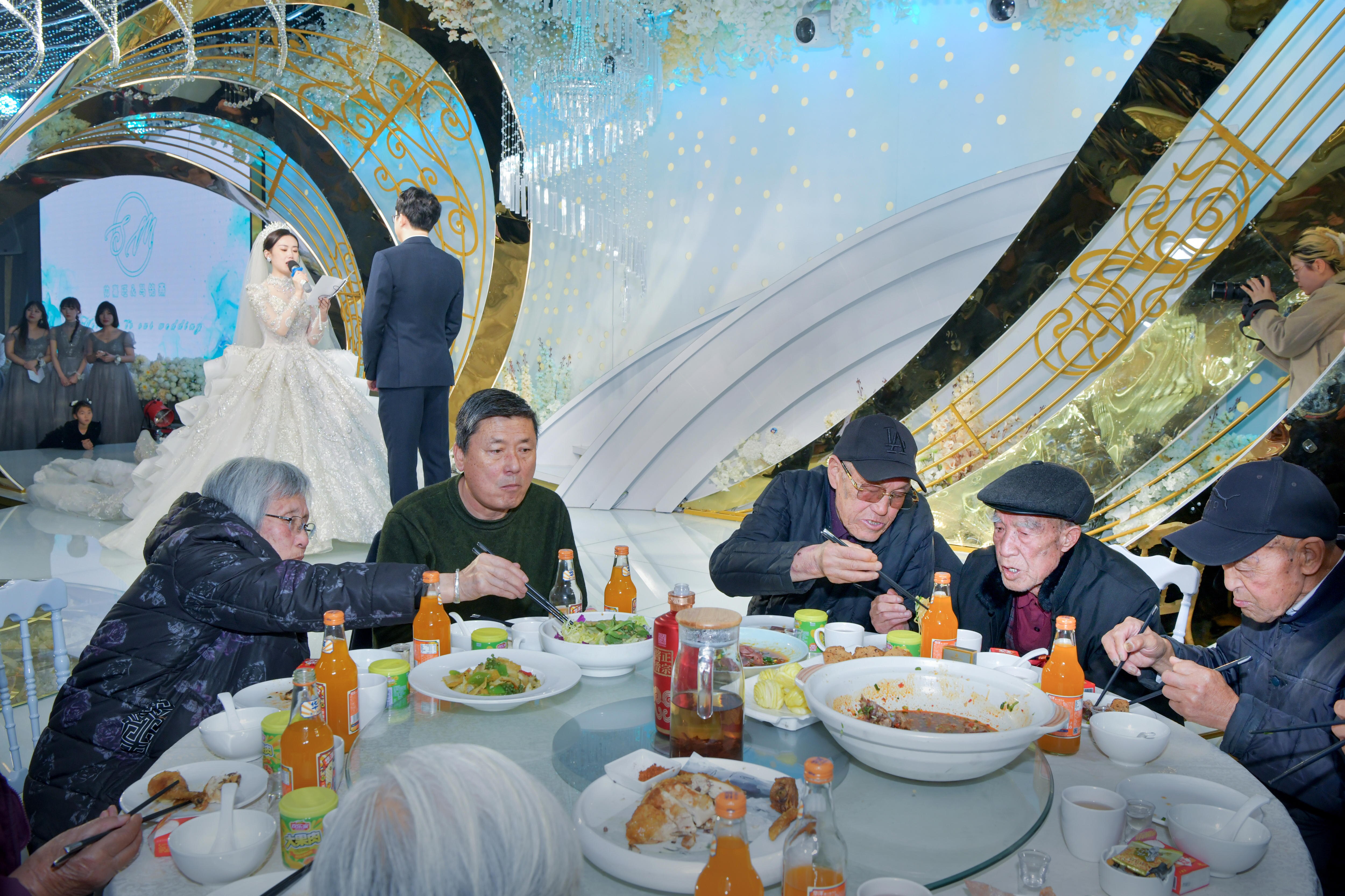 A group of elderly people sit at a round table, sharing food. A bride and groom stand in the background, giving speeches