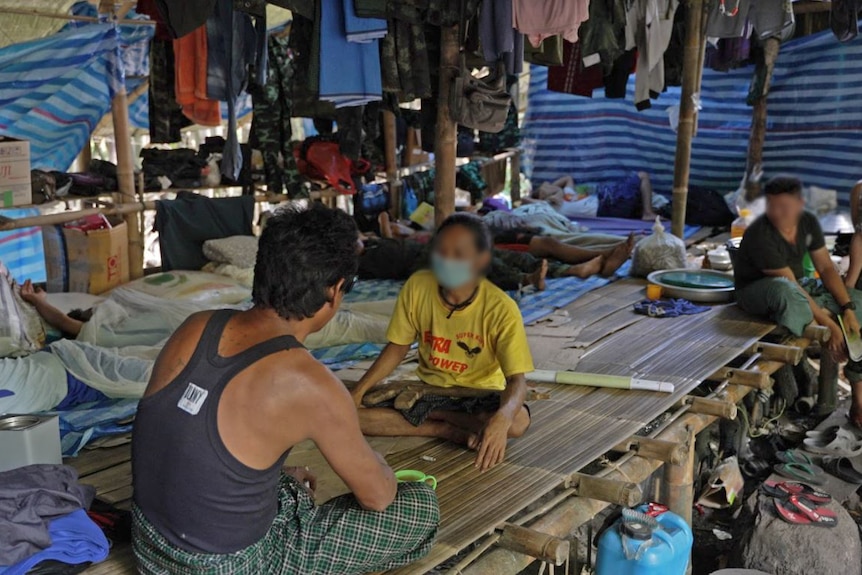 A man sits on a board facing another person wearing a yellow shirt and mask.