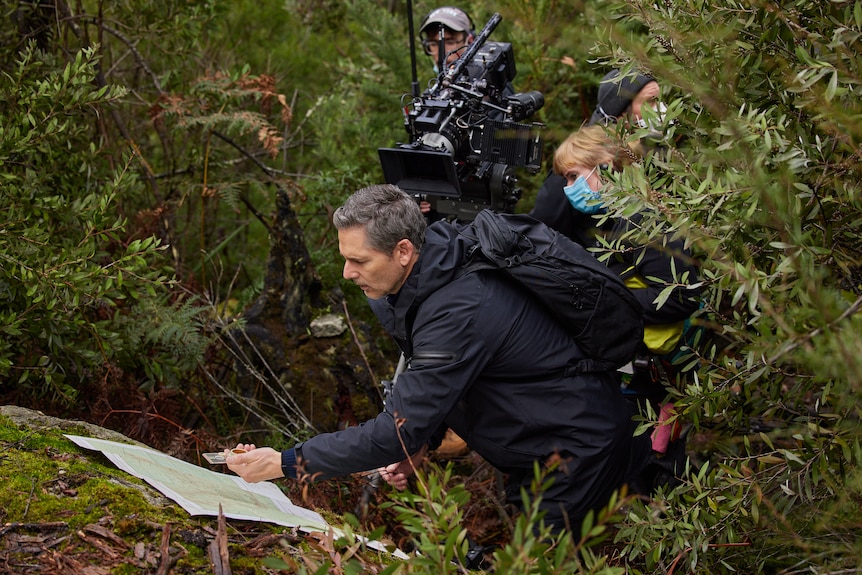 Actor Eric Bana pointing at a map as a film crew shoots behind him, surrounded by bushland