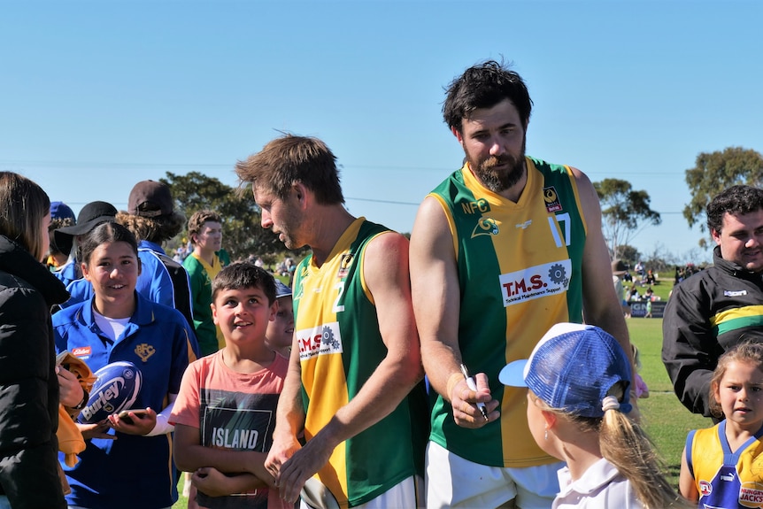 Two men in green and gold football uniforms hold pens to sign autographs for young fans looking up at them 