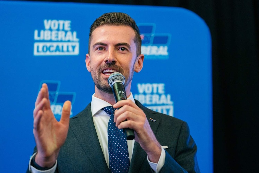 A close up of Zak Kirkup on stage with a microphone in front of a blue Liberal Party banner.