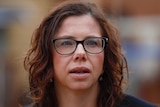 A woman with brown curly hair and glasses mid-sentence with everything else around her blurred out