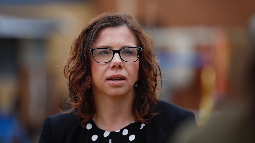 A woman with brown curly hair and glasses mid-sentence with everything else around her blurred out