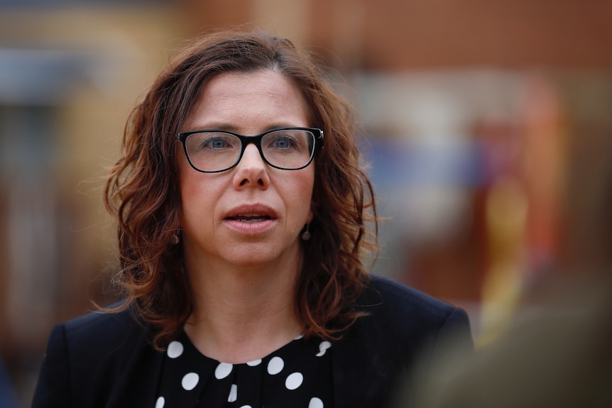 A woman with brown curly hair and glasses mid-sentence with everything else around her blurred out