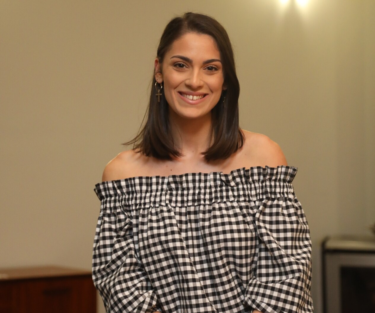 Photograph of a young woman smiling at the camera. She has a black and white gingham off the shoulders top, and dark hair