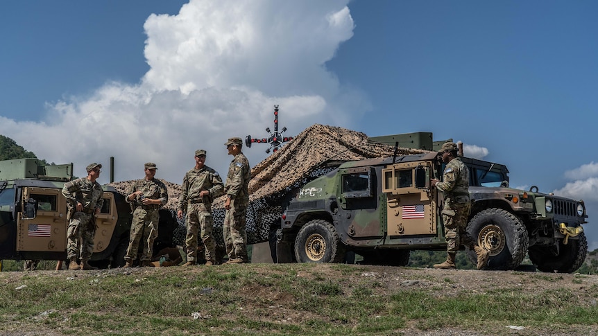 A group of soldiers dressed in army fatigues stand beside an army car on a base.