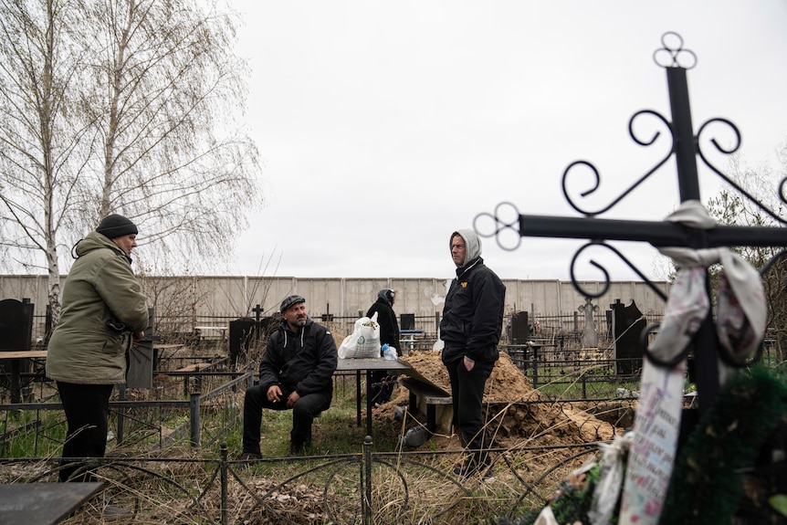 Four men hanging around a graveyard, two with cigarettes, on an overcast day