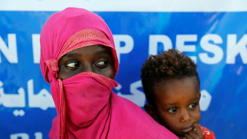 A woman wears a pink headscarf as she stands with her young child at a refugee camp