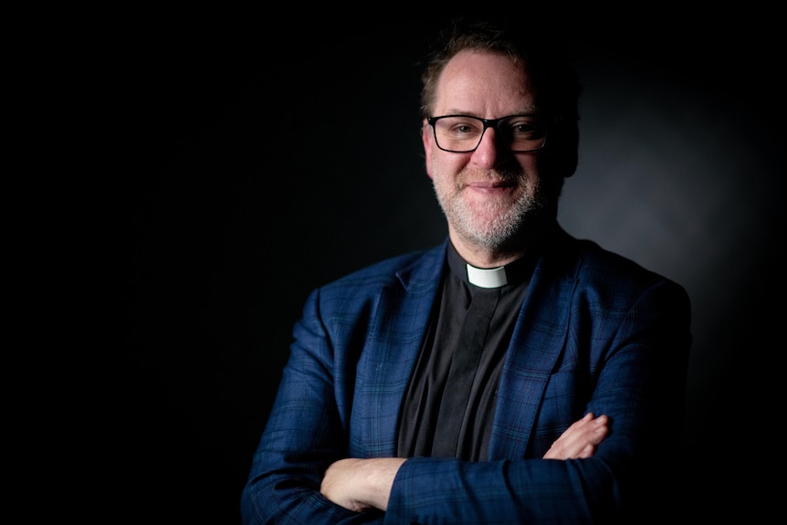  A man wearing a clerical collar and a black shirt smiles at the camera while crossing his arms.