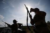 Two men with rifles train at shooting range, with one inspecting his gun while the other reloads.