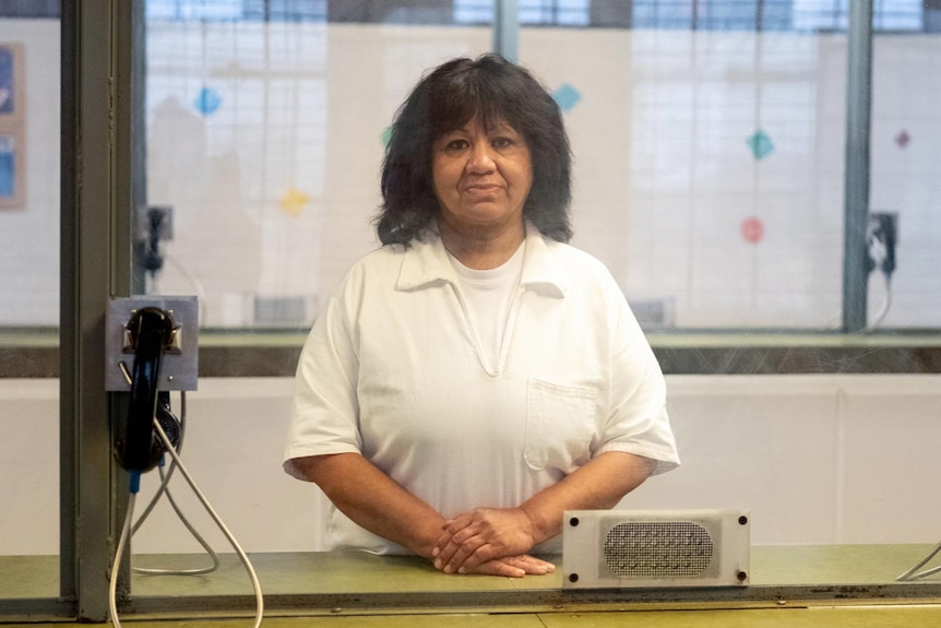 An older woman in prison carb stands behind glass with hands resting in front of her.